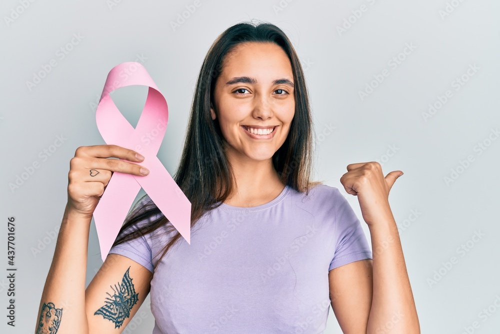 Young hispanic woman holding pink cancer ribbon pointing thumb up to the side smiling happy with open mouth Young hispanic woman holding pink cancer ribbon pointing thumb up to the side smiling happy with open mouth
