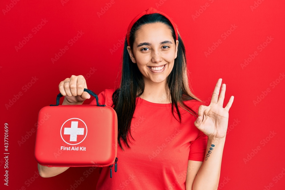 Young hispanic woman holding first aid kit doing ok sign with fingers, smiling friendly gesturing excellent symbol Young hispanic woman holding first aid kit doing ok sign with fingers, smiling friendly gesturing excellent symbol