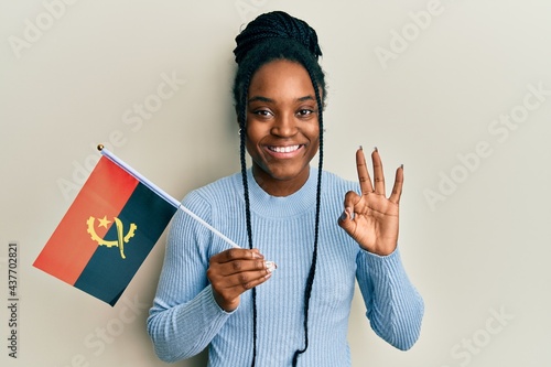 African american woman with braided hair holding angola flag doing ok sign with fingers, smiling friendly gesturing excellent symbol photo