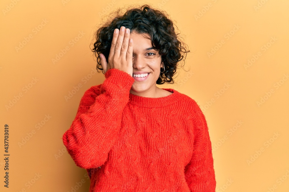 Young hispanic woman with curly hair wearing casual winter sweater covering one eye with hand, confident smile on face and surprise emotion. Young hispanic woman with curly hair wearing casual winter sweater covering one eye with hand, confident smile on face and surprise emotion.