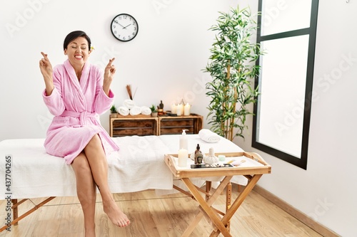 Beautiful caucasian woman wearing bathrobe at wellness spa gesturing finger crossed smiling with hope and eyes closed. luck and superstitious concept.