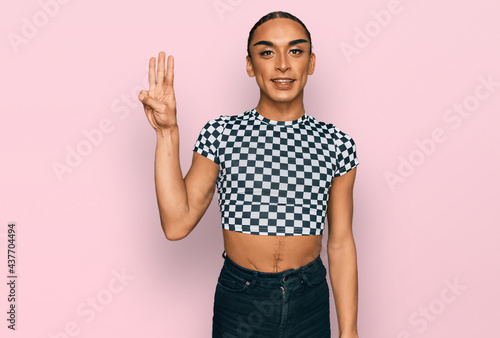 Hispanic transgender man wearing make up and long hair wearing modern clothes showing and pointing up with fingers number three while smiling confident and happy.