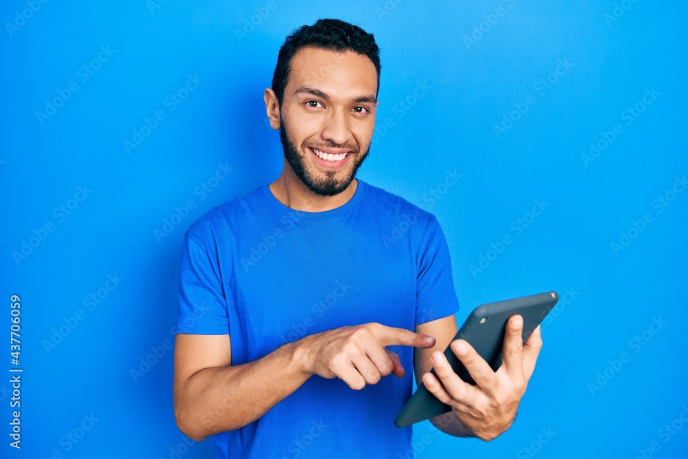 Hispanic man with beard using touchpad device smiling happy pointing with hand and finger Hispanic man with beard using touchpad device smiling happy pointing with hand and finger