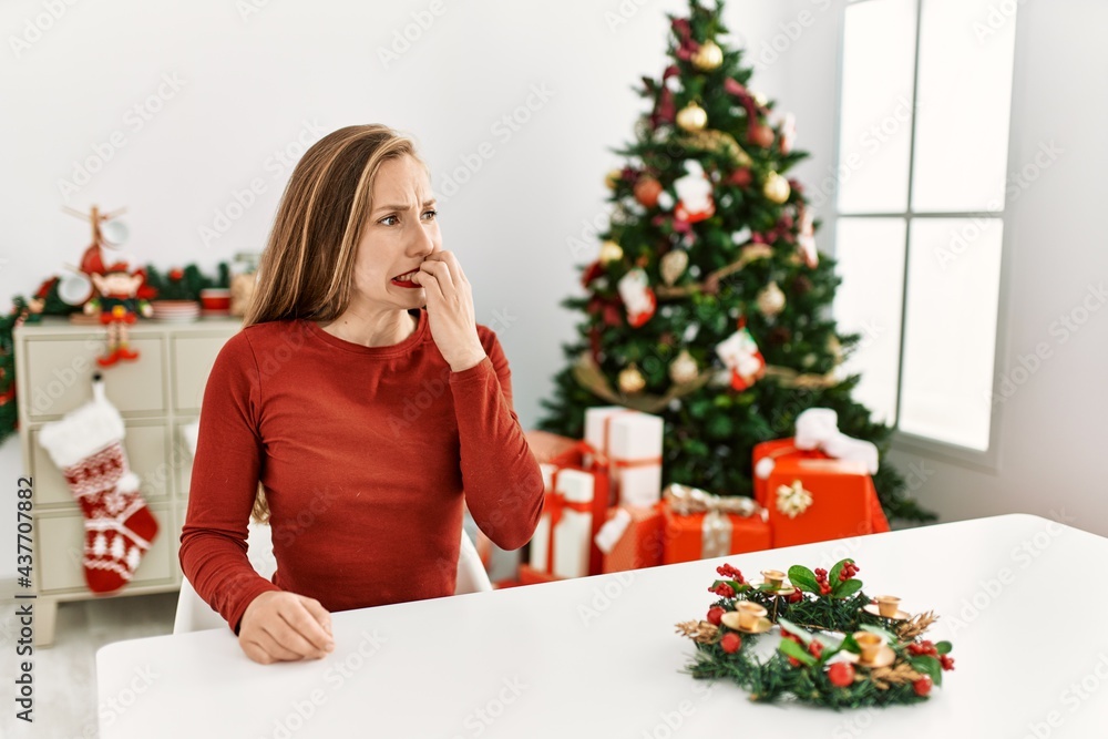 Caucasian young blonde woman sitting on the table by christmas tree looking stressed and nervous with hands on mouth biting nails. anxiety problem. Caucasian young blonde woman sitting on the table by christmas tree looking stressed and nervous with hands on mouth biting nails. anxiety problem.