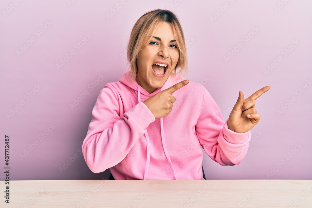 Fototapeta premium Young caucasian woman wearing casual clothes sitting on the table smiling and looking at the camera pointing with two hands and fingers to the side.