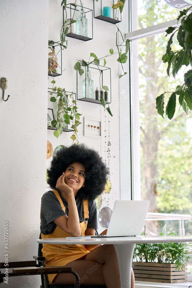 Young thoughtful dreamy smiling African American student generation z girl with afro hair sitting at table with laptop thinking about inspiration ideas in modern cafe. - obrazy, fototapety, plakaty 