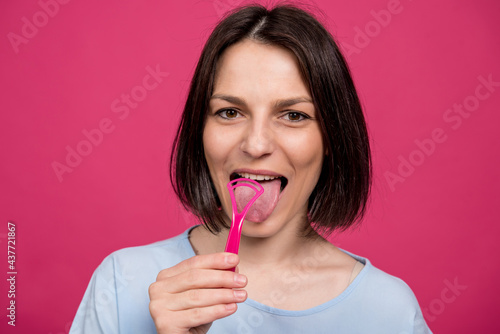 Beautiful happy young woman with tongue scraper on blank pink background