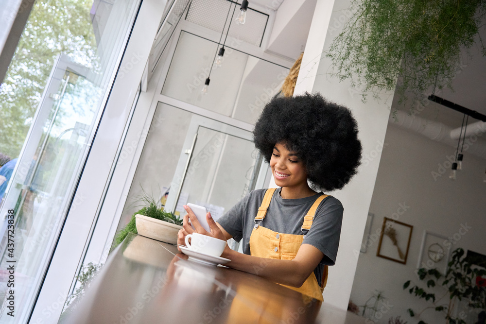 Young happy gen z black student hipster girl with afro hair sitting at table in cafe indoor using mobile shop marketing app online watching video working typing surfing internet. - obrazy, fototapety, plakaty 