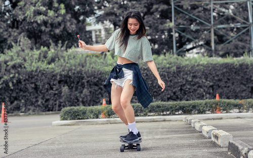 Beautiful happy Asian healthy woman smiling, holding sweet lollipop, riding and playing extreme sportive skateboard as outdoor activity with happiness, relaxation during holidays in summer vacation.