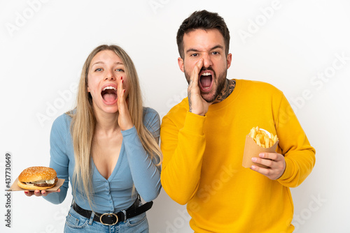 Couple holding hamburger and fried chips over isolated white background shouting and announcing something