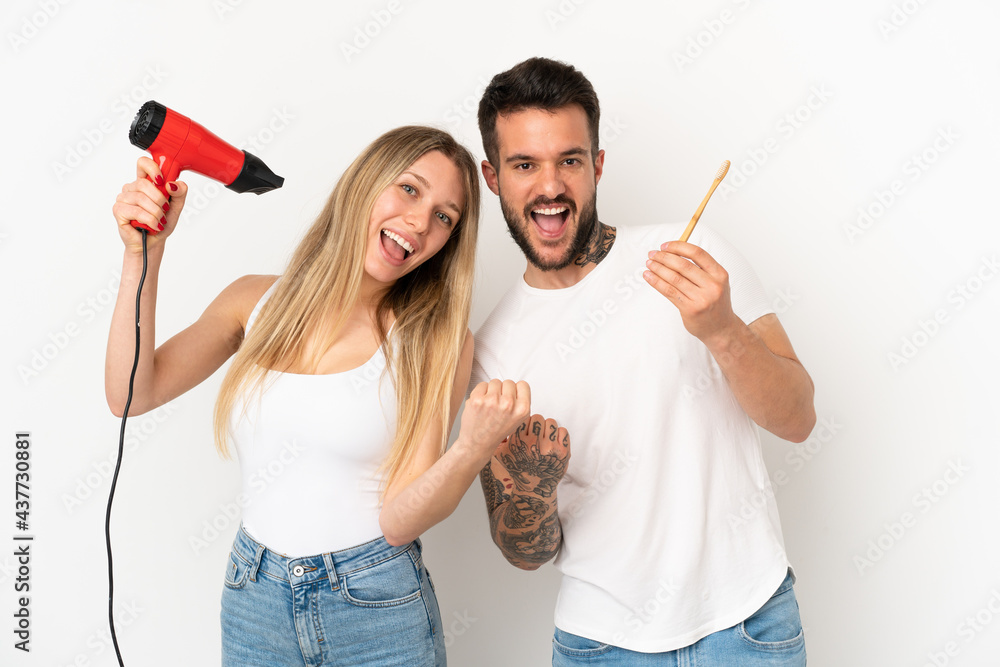 Couple holding a hairdryer and brushing teeth over isolated white background celebrating a victory Couple holding a hairdryer and brushing teeth over isolated white background celebrating a victory