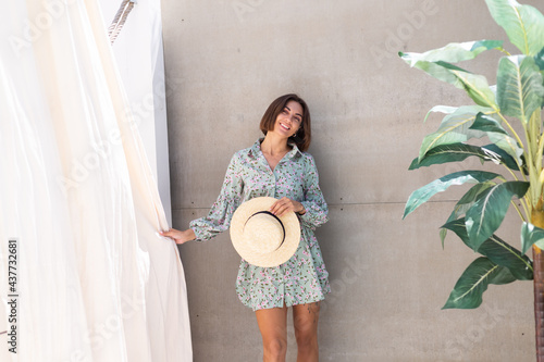 Beautiful woman in summer dress and straw hat by palm tree and gray wall on background, playful joyful positive excited cheerful happy