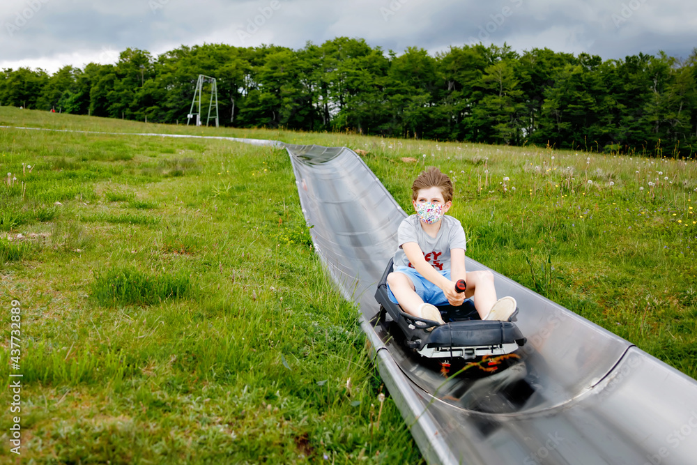 Young school kid boy having fun riding summer toboggan run sled down a hill in Hoherodskopf, Germany. Active child with medical mask making funny activity otudoors. Family leisure with kids. Young school kid boy having fun riding summer toboggan run sled down a hill in Hoherodskopf, Germany. Active child with medical mask making funny activity otudoors. Family leisure with kids.