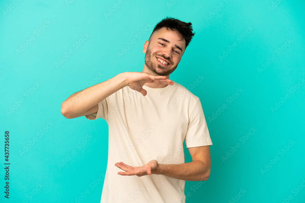 Young caucasian man isolated on blue background holding copyspace imaginary on the palm to insert an ad Young caucasian man isolated on blue background holding copyspace imaginary on the palm to insert an ad