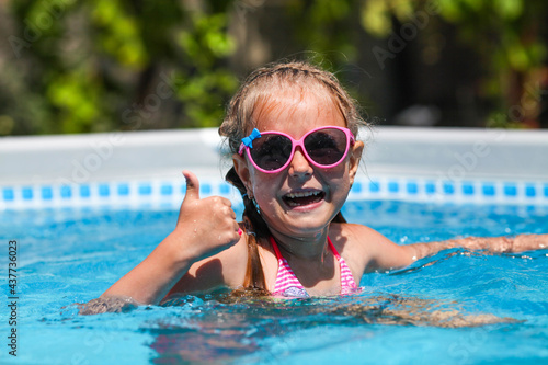 Cute little girl smiling in sunglasses in the pool on a sunny day. The child showing thumbs up like gesture looking camera . Summer vacation