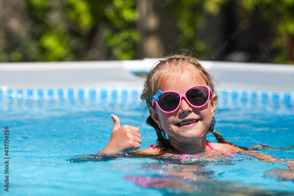 Cute little girl smiling in sunglasses in the pool on a sunny day. The child showing thumbs up like gesture looking camera . Summer vacation