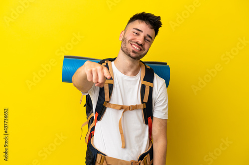 Young mountaineer caucasian man with a big backpack isolated on yellow background pointing front with happy expression