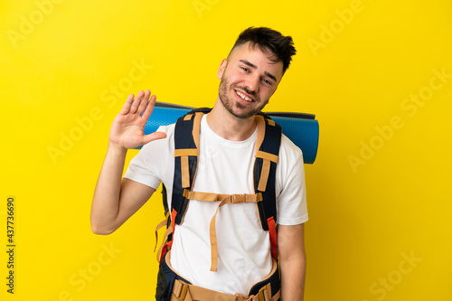 Young mountaineer caucasian man with a big backpack isolated on yellow background saluting with hand with happy expression