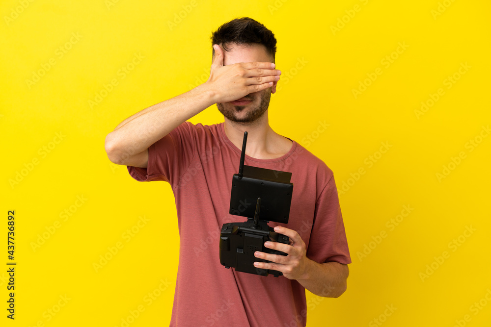 Man holding a drone remote control isolated on yellow background covering eyes by hands. Do not want to see something Man holding a drone remote control isolated on yellow background covering eyes by hands. Do not want to see something