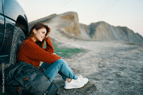 woman in jeans and sneakers sits on the sand near the car in nature