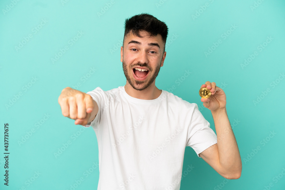 Young man holding a Bitcoin isolated on blue background surprised and pointing front Young man holding a Bitcoin isolated on blue background surprised and pointing front