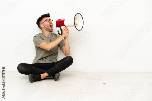 Young caucasian man sitting on the floor isolated on white background shouting through a megaphone