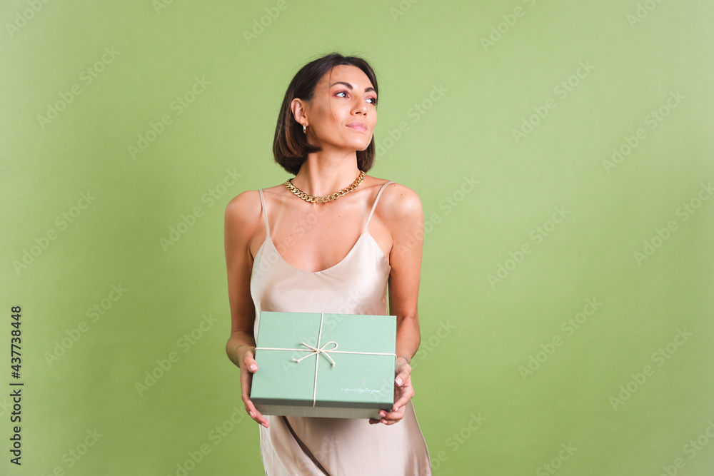 Fototapeta premium Woman in silk golden dress on green background holding gift box happy positive excited cheerful posing