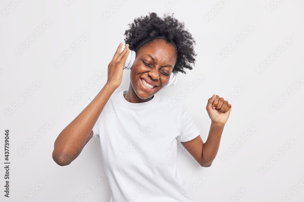 Studio shot of upbeat dark skinned young woman chills while listens music in headphones dances and catches every bit smiles broadly has snow white teeth dressed casually. Enjoying favorite song Studio shot of upbeat dark skinned young woman chills while listens music in headphones dances and catches every bit smiles broadly has snow white teeth dressed casually. Enjoying favorite song