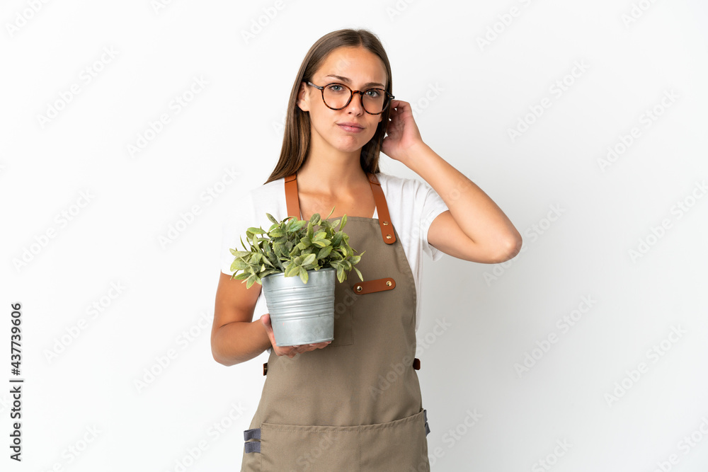 Gardener girl holding a plant over isolated white background having doubts Gardener girl holding a plant over isolated white background having doubts