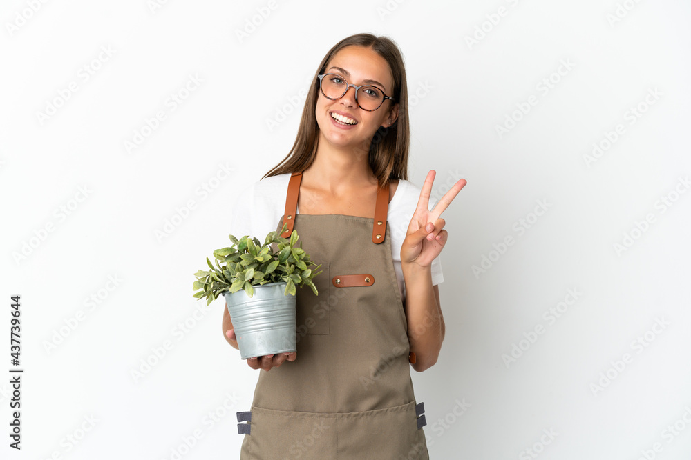 Gardener girl holding a plant over isolated white background smiling and showing victory sign Gardener girl holding a plant over isolated white background smiling and showing victory sign