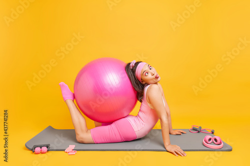 Fototapeta Naklejka Na Ścianę i Meble -  Funny sporty woman foolishes around whle doing pilates exercises makes grimace trains with fitball poses on mat surrounded by sport equipment stays fit. Active lifestyle and training concept