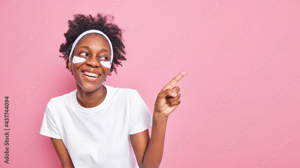 Happy young Afro American woman with curly hair smiles broadly shows white perfect teeth applies patches under eyes dressed in casual t shirt indicates on blank space against pink background Happy young Afro American woman with curly hair smiles broadly shows white perfect teeth applies patches under eyes dressed in casual t shirt indicates on blank space against pink background