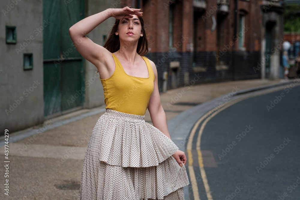 Young woman in a yellow t-shirt and white skirt posing in a narrow street.