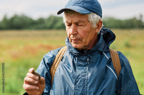 Closeup portrait of treasure hunter man wearing cap and jacket holding coin found in ground in hands and looking with concentrated facial expression at his finding.