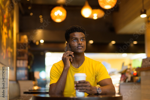 African man wearing yellow t-shirt in coffee shop while talking on mobile phone
