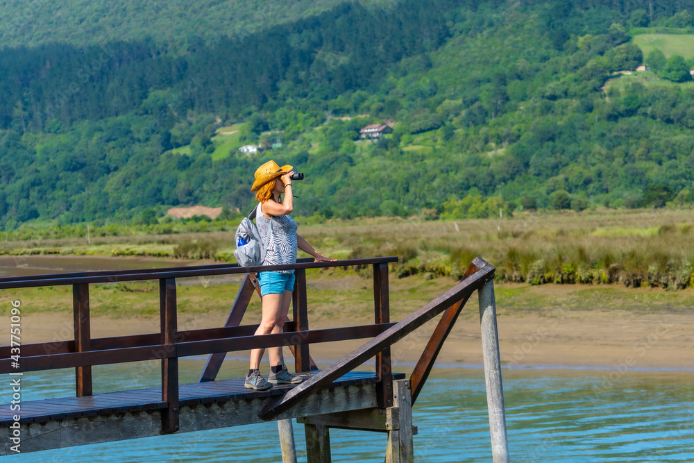 Naklejka premium A young woman with spyglasses looking at the birds from the wooden piers of the Urdaibai marshes, a biosphere reserve in Bizkaia next to Mundaka. Basque Country