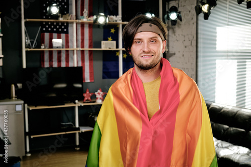 Happy young man wrapped into pride flag standing in living-room