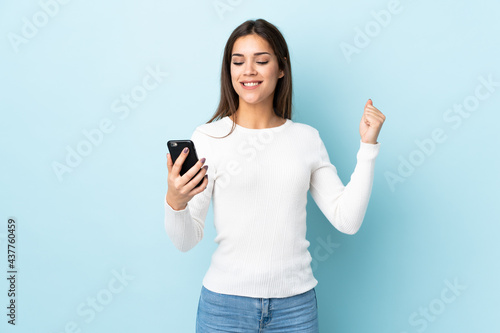 Young caucasian woman isolated on blue background with phone in victory position