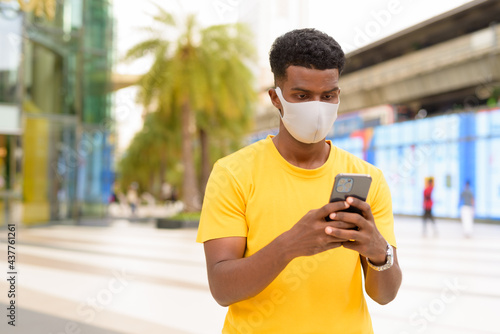 African man wearing yellow t-shirt and face mask to protect from Covid-19 coronavirus outdoors in city while using mobile phone