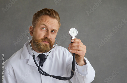 Headshot portrait handsome male doctor with funny pensive face expression holding stethoscope. Serious doc in white coat listening to patient's breath or examining heartbeat. Medical checkup concept
