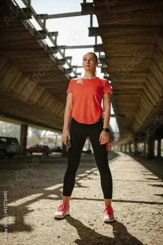 Woman posing full length wearing sportswear under the bridge, looking at camera and holding phone.