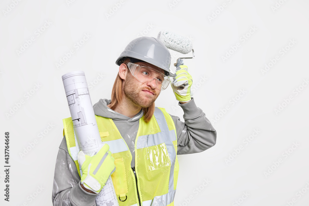 Frustrated displeased thoughtful male builder holds painting roller and blueprint busy doing home improvement dressed in uniform protective hardhat isolated over white background blank space Frustrated displeased thoughtful male builder holds painting roller and blueprint busy doing home improvement dressed in uniform protective hardhat isolated over white background blank space