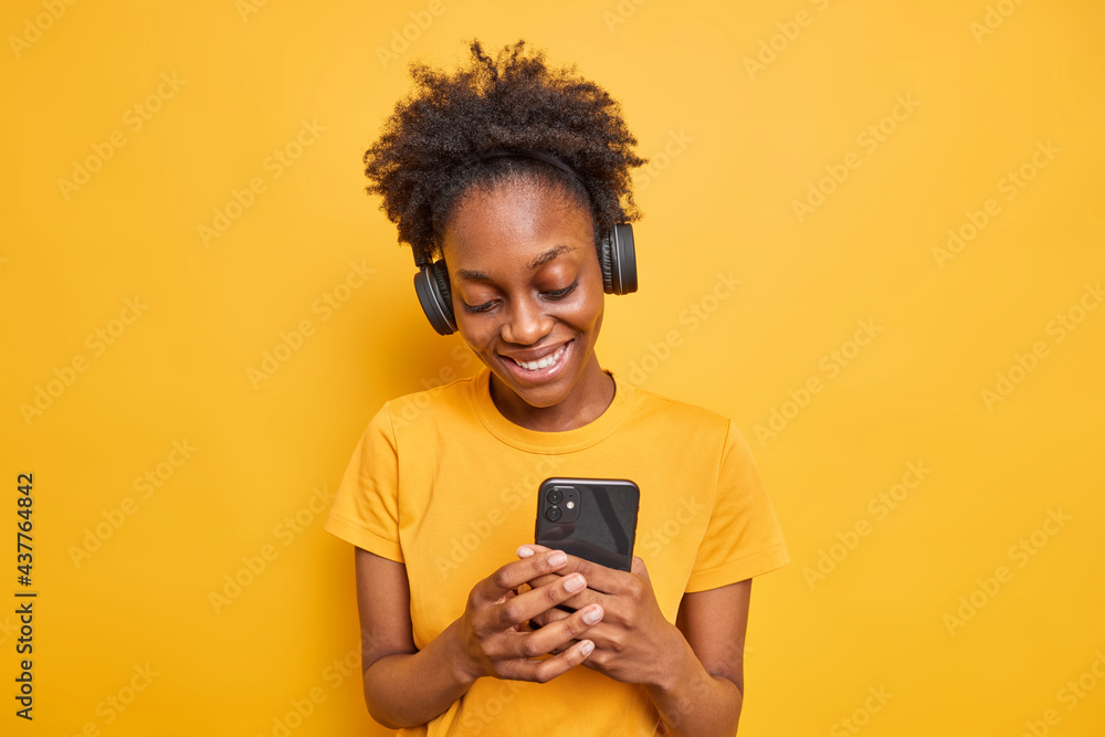 Studio shot of smiling teenage girl with dark skin sends text messages via smartphone chooses song from playlist listens music wears stereo headphones on ears isolated over yellow background