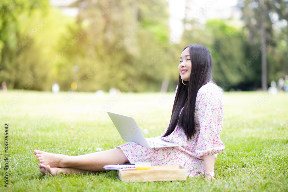 Working outside in summer or spring and WFH concept, Young beautiful Asian woman reading a book and looking laptop in the natural park, Freelance working, homeschooling, distance learning.