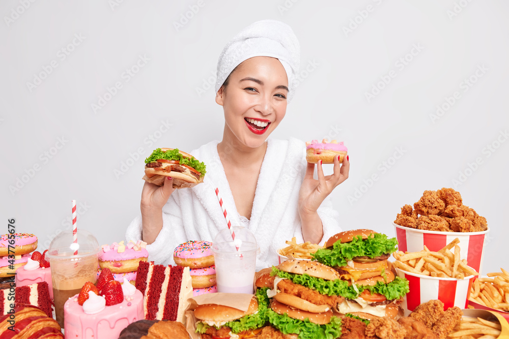 Hungry Asian lady smiles gladfully holds doughnut and sandwich surrounded by fast food dressed in bathrobe shower towel over head after taking bath isolated over white background. Cheat meal Hungry Asian lady smiles gladfully holds doughnut and sandwich surrounded by fast food dressed in bathrobe shower towel over head after taking bath isolated over white background. Cheat meal