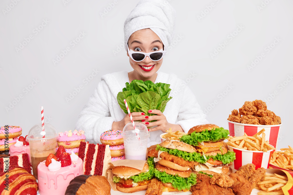 Positive Asian woman with healthy skin wears sunglasses domesticrobe and towel on head holds green vegetable avoids eating junk food which surround her chooses healthy food and proper nutrition Positive Asian woman with healthy skin wears sunglasses domesticrobe and towel on head holds green vegetable avoids eating junk food which surround her chooses healthy food and proper nutrition