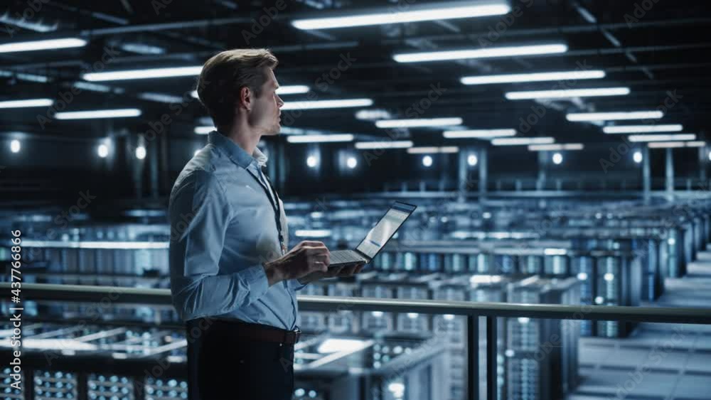 Portrait of IT Specialist Uses Laptop in Data Center. Server Farm Cloud Computing Facility with Male Maintenance Administrator Working. Cyber Security and Network Protection. Medium Tracking Wide Portrait of IT Specialist Uses Laptop in Data Center. Server Farm Cloud Computing Facility with Male Maintenance Administrator Working. Cyber Security and Network Protection. Medium Tracking Wide