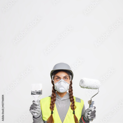 Vertical shot of serious woman wears protective helmet glasses and respirator holds repair tools focused above going to redecorate apartment poses against white background blank space overhead
