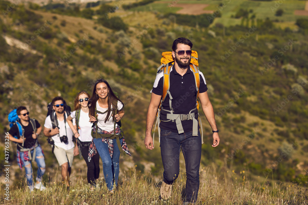 Group of smiling young travelers hikers tourists with backpacks hiking with in row in green valley during summer vacations on sunny clear day. Hiking and traveling together, active lifestyle