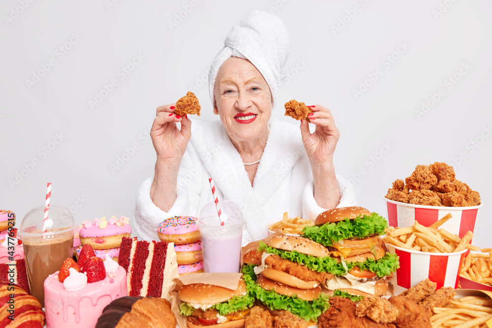 Bad eating habits. Happy wrinkled old woman eats junk food has obsession to cheat meal wears soft white dressing gown and towel over head poses against white background with delicious snacks around Bad eating habits. Happy wrinkled old woman eats junk food has obsession to cheat meal wears soft white dressing gown and towel over head poses against white background with delicious snacks around